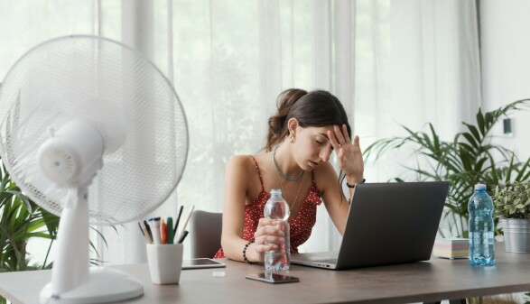 Eine junge Frau arbeitet bei Hitze zu Hause am Schreibtisch und hat einen Ventilator laufen.
