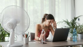Eine junge Frau arbeitet bei Hitze zu Hause am Schreibtisch und hat einen Ventilator laufen.