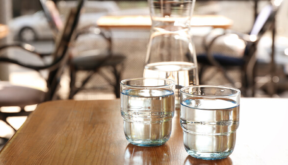Zwei Gl&auml;ser Wasser auf einem Tisch in einem Restaurant. Dahinter ist eine Karaffe mit Wasser zu sehen.