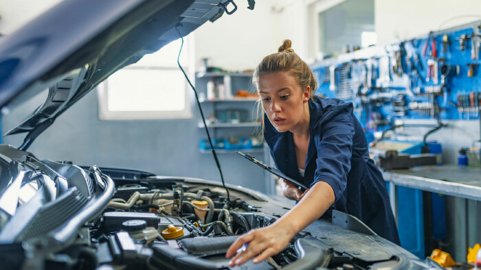 Mechanikerin in blauer Arbeitskleidung untersucht den Motorraum eines Autos in einer Werkstatt.