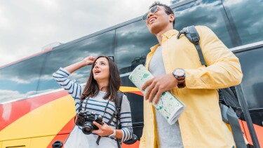 Eine Frau mit Digitalkamera blickt zur Seite, w&auml;hrend ein Mann unterschiedlicher ethnischer Herkunft neben einem Reisebus eine Karte h&auml;lt.