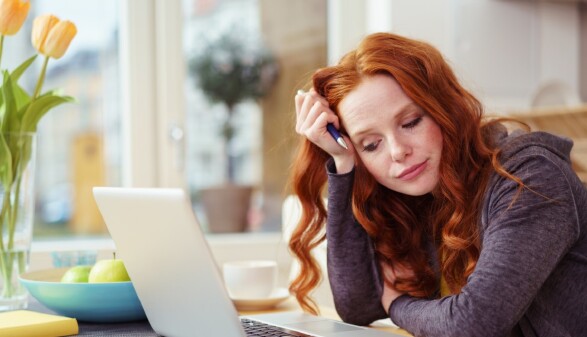 Junge Frau sitzt am K&uuml;chentisch und schaut auf ihren Laptop.