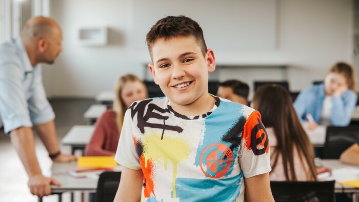 Sch&uuml;ler sitzt l&auml;chelnd in der Klasse und blickt in Richtung Kamera