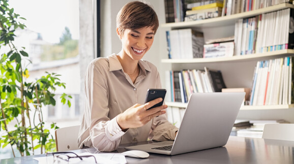 Frau sitzt lachend am Schreibtisch mit einem Smartphone in der Hand. Vor ihr steht ein Notebook.