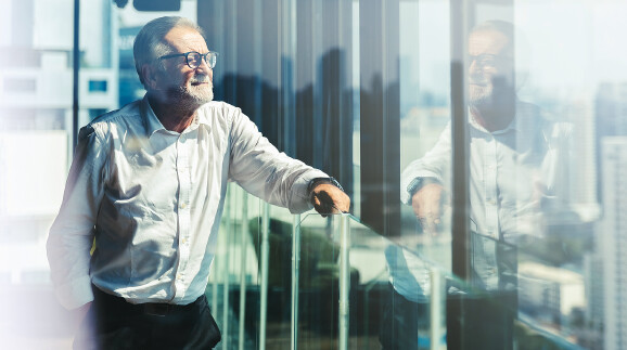 Ein &auml;lterer Gesch&auml;ftsmann steht in einer modernen Lounge hoch oben in einem B&uuml;roturm und blickt nach rechts.
