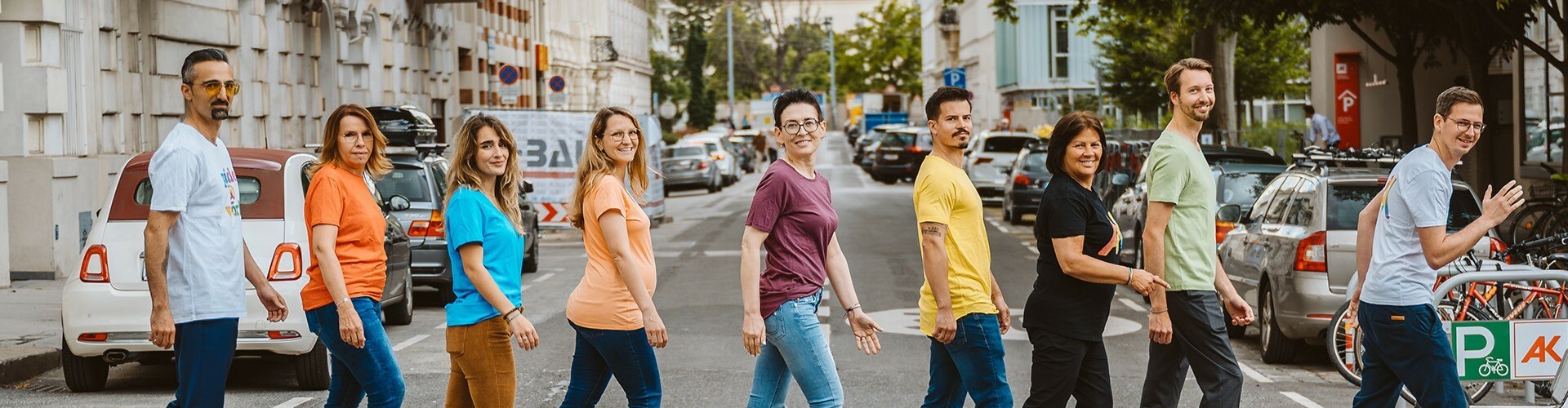 AK Präsidentin Renate Anderl und 8 Mitarbeiter:innen der AK Wien gehen in bunten T-Shirts über einen Zebrastreifen und lachen in die Kamera