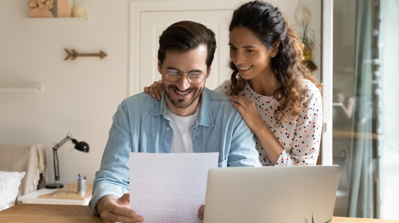 Mann sitzt am Schreibtisch und schaut freudig auf ein Blatt Papier, Frau umarmt ihn von hinten, liest mit und l&auml;chelt