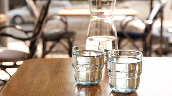 Zwei Gl&auml;ser Wasser auf einem Tisch in einem Restaurant. Dahinter ist eine Karaffe mit Wasser zu sehen.