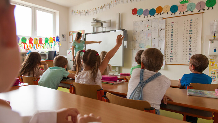 Lehrerin zeigt auf eine interaktive Tafel in einer Volksschule. Volkssch&uuml;ler:innen lernen und eine Sch&uuml;lerin meldet sich.