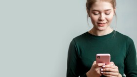 Nette junge Frau trägt ein grünes langärmeliges Hemd und hält ein Mobiltelefon in der Hand.