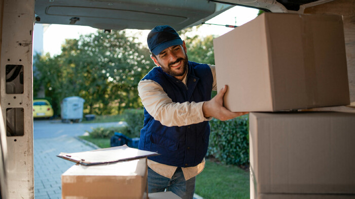Ein Paketzusteller lädt bei Tageslicht Kartons aus einem Lieferwagen und lächelt dabei freundlich.