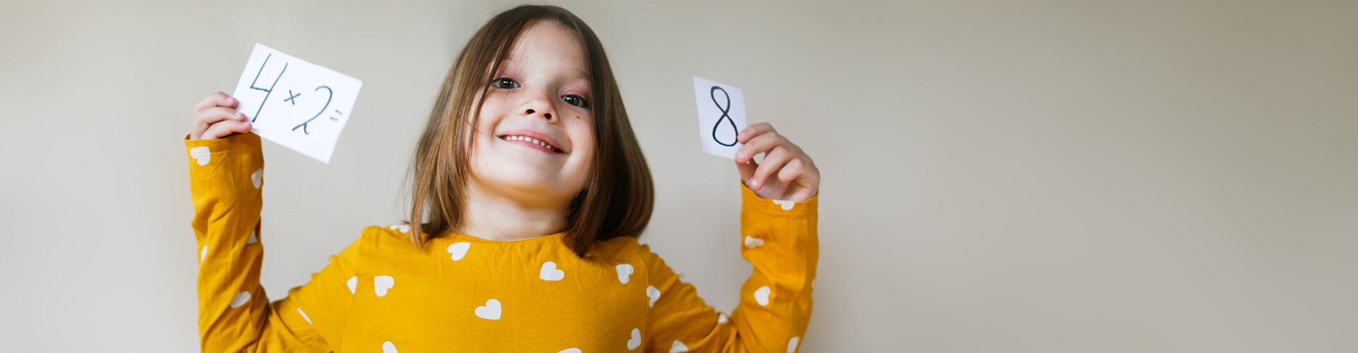 Mädchen im Volksschulalter lacht in die Kamera und hält ein Rechenbeispiel auf Zetteln hoch