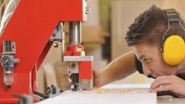 Ein männlicher Jugendlicher mit Gehörschutz steht an einer Werkbank und bearbeitet ein Holzbrett mit einer Fräsmaschine. © jörn buchheim, stock.adobe.com