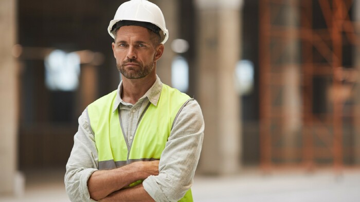  Portr&auml;t eines reifen Bauarbeiters, der in die Kamera blickt, w&auml;hrend er mit verschr&auml;nkten Armen auf der Baustelle steht.