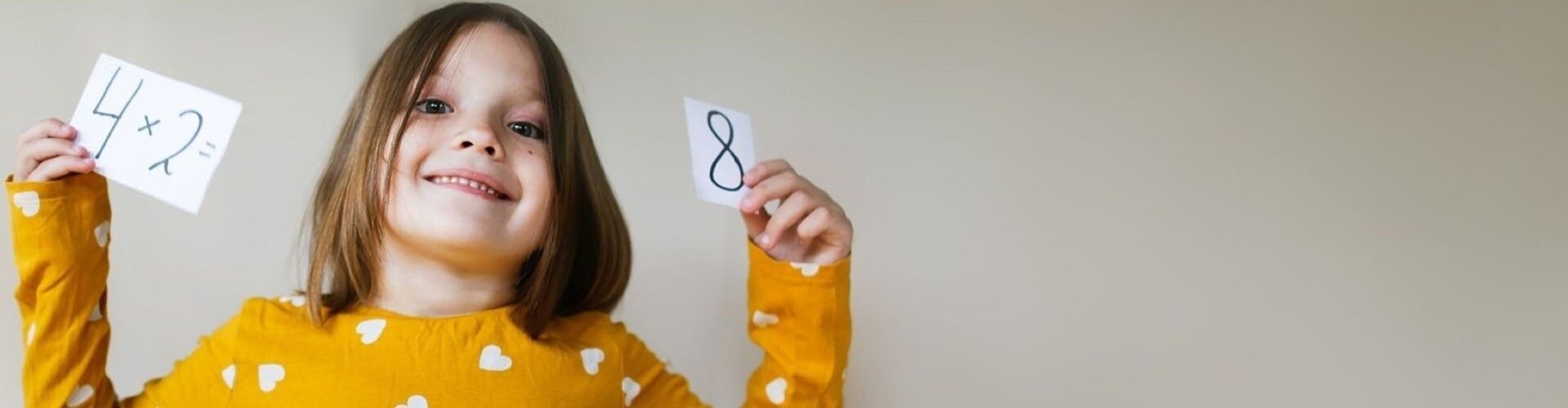 Mädchen im Volksschulalter lacht in die Kamera und hält ein Rechenbeispiel auf Zetteln hoch