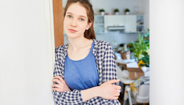Junge Frau steht im Türrahmen einer geöffnete Zimmertüre und schaut mit verschränkten Armen in die Kamera. Hinter ihr sieht man unscharf eine Küche und geöffnete Kartons auf einem Tisch stehen.