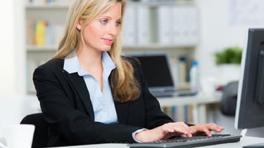 Eine Frau im Anzug sitzt am Schreibtisch und blickt auf den Computerbildschirm vor sich. Im Hintergrund sieht man ein Regal mit Ordnern. © Racle Fotodesign, stock.adobe.com
