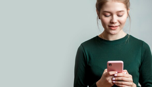 Nette junge Frau trägt ein grünes langärmeliges Hemd und hält ein Mobiltelefon in der Hand.