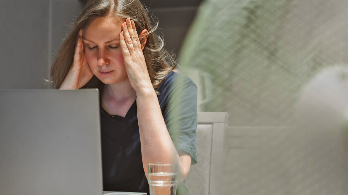 Frau sitzt vor einem Laptop am Schreibtisch, vor ihr ein Ventilator, und h&auml;lt sich mit den H&auml;nden den Kopf, hat ersch&ouml;pft die Augen geschlossen